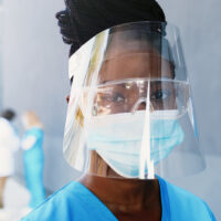 Portrait African American beautiful woman doctor in medical mask, face shield and goggles looking at camera. Close up female physician in respiratory protection. Doctors on background. Protected suit.